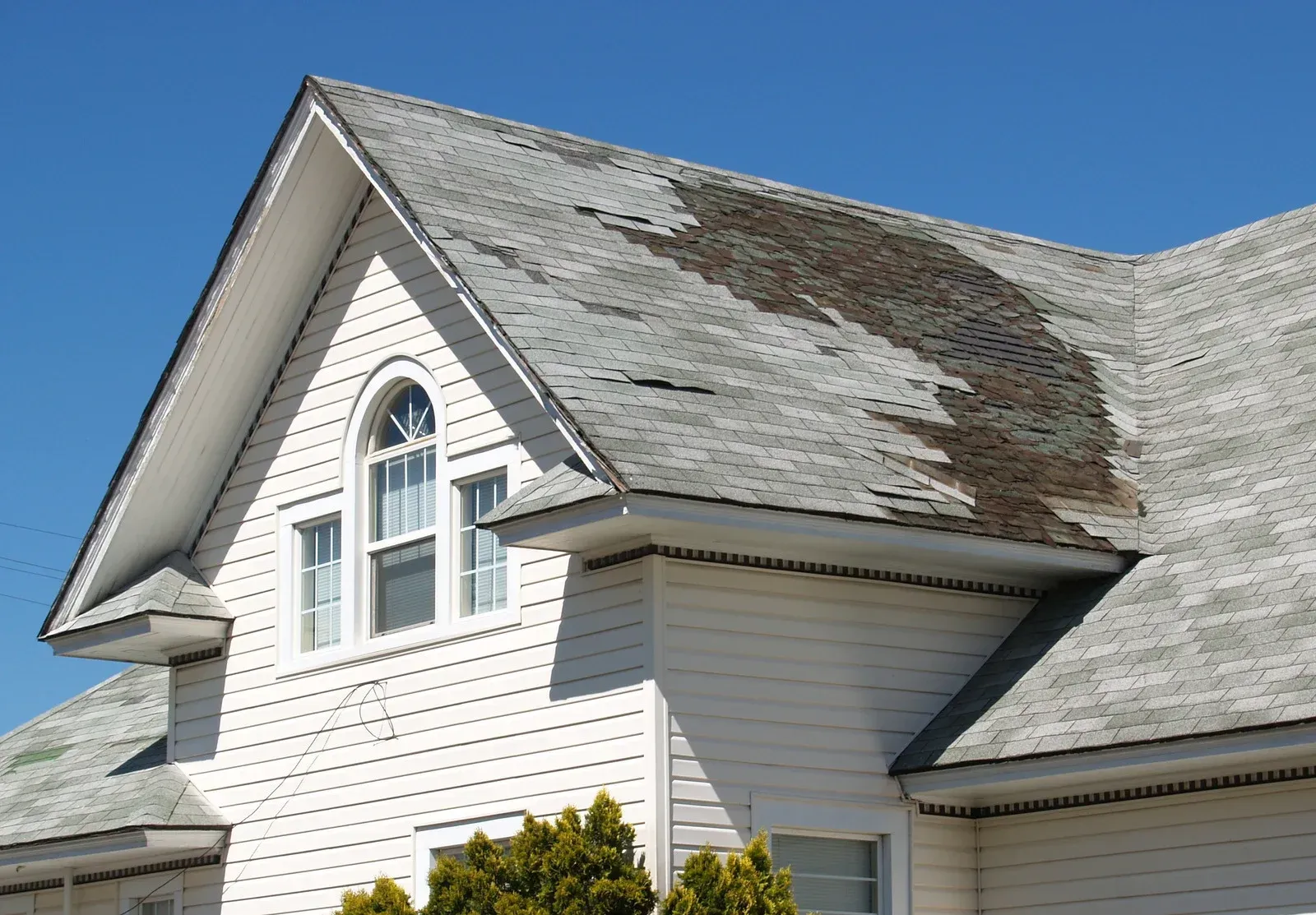 White house roof with extensive dark weather damage and peeling shingles against a blue sky