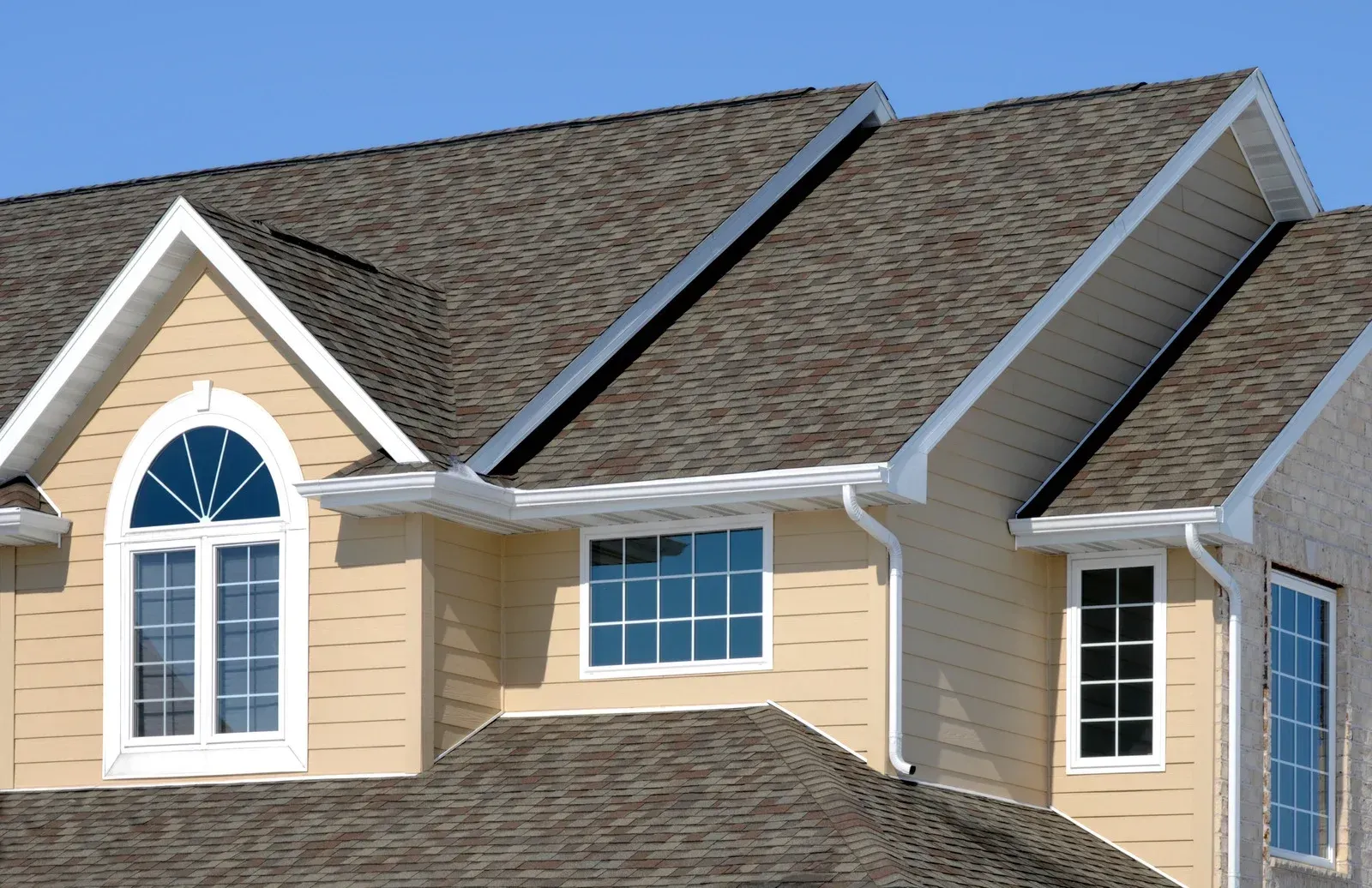 Suburban house exterior with beige siding, white trim, and dark shingled roof under a blue sky