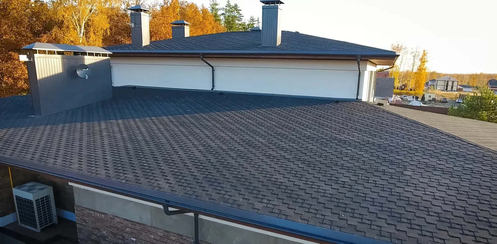 Rooftop with brown shingle tiles and white stucco walls under a clear sky, with autumn trees in the background
