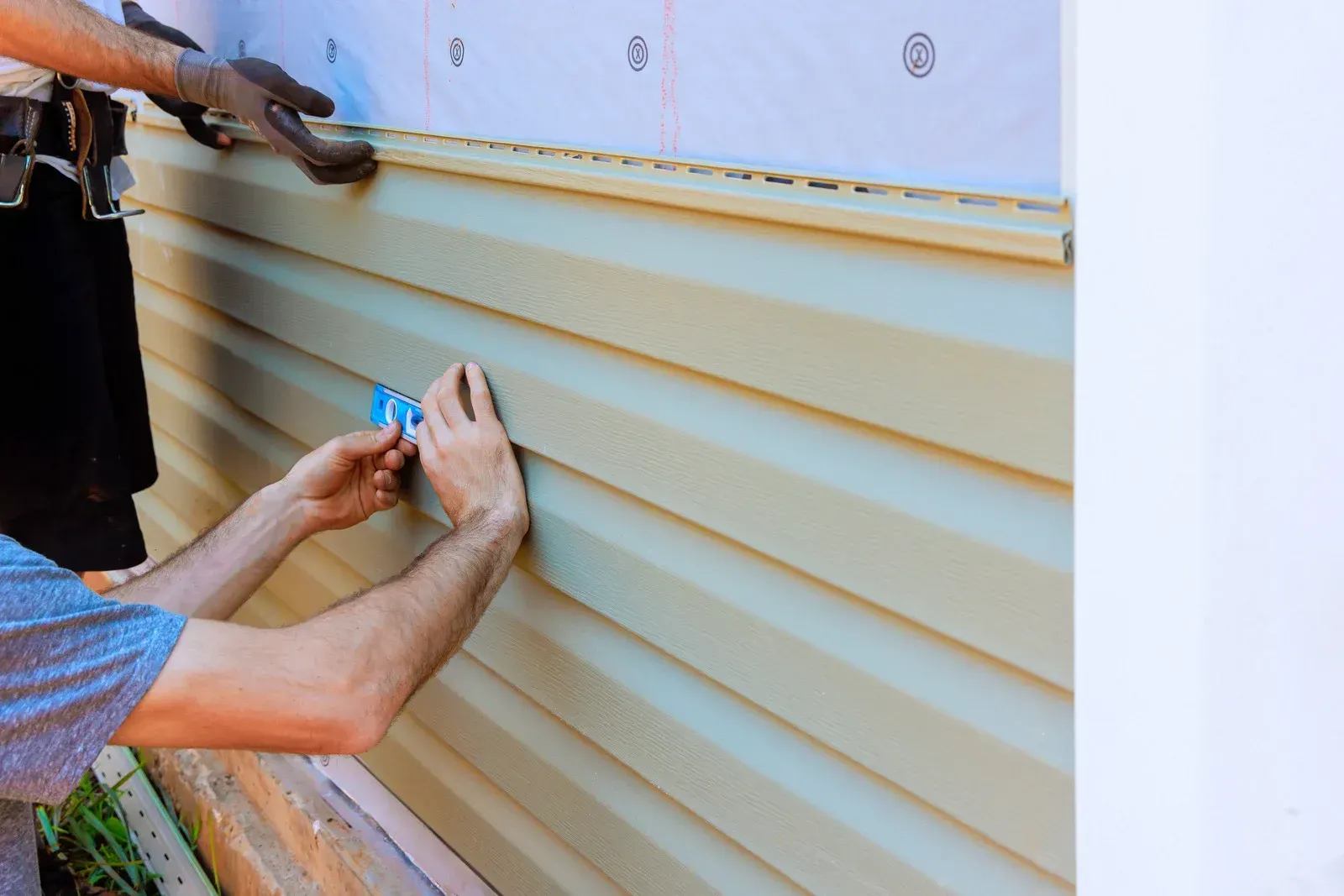 Two workers install beige vinyl siding on a house exterior using hand tools.