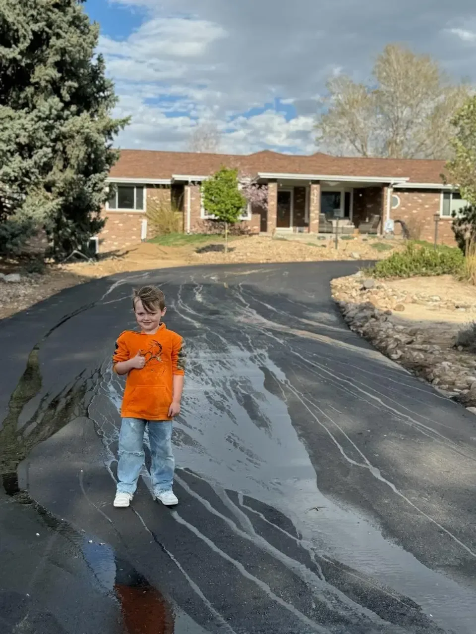 A young boy in an orange shirt is standing in a puddle of water in front of a house