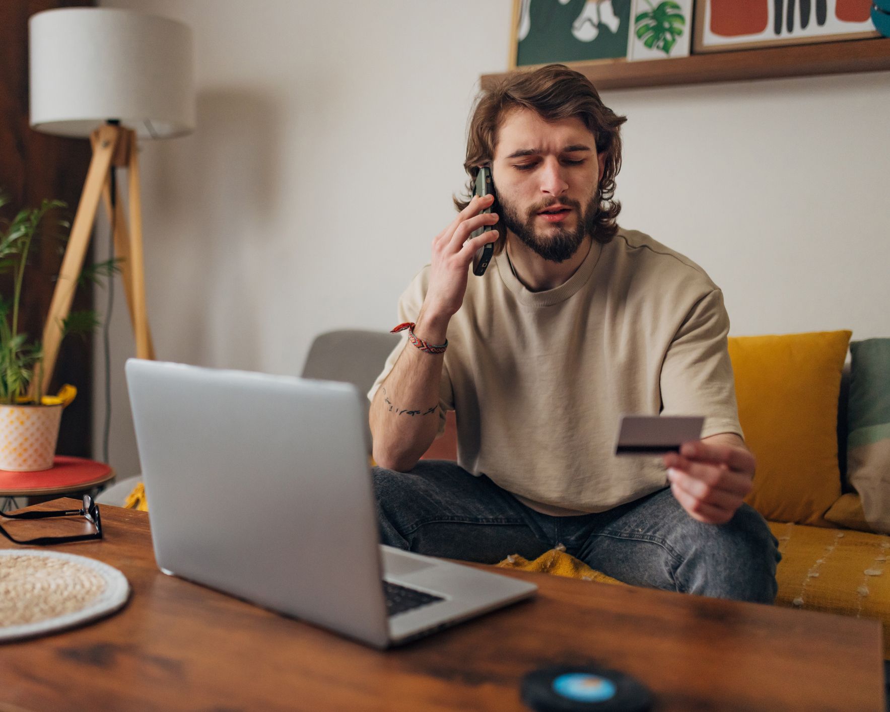 A man is sitting on a couch holding a credit card and talking on a cell phone.