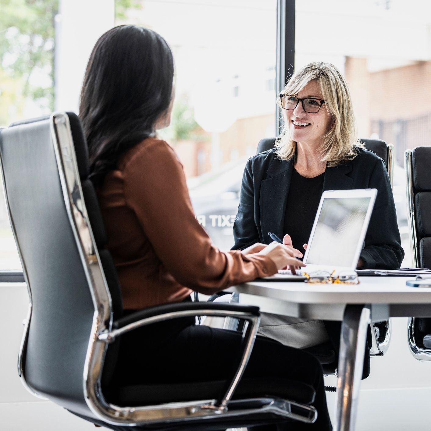 Two women are sitting at a table with a laptop and talking to each other.