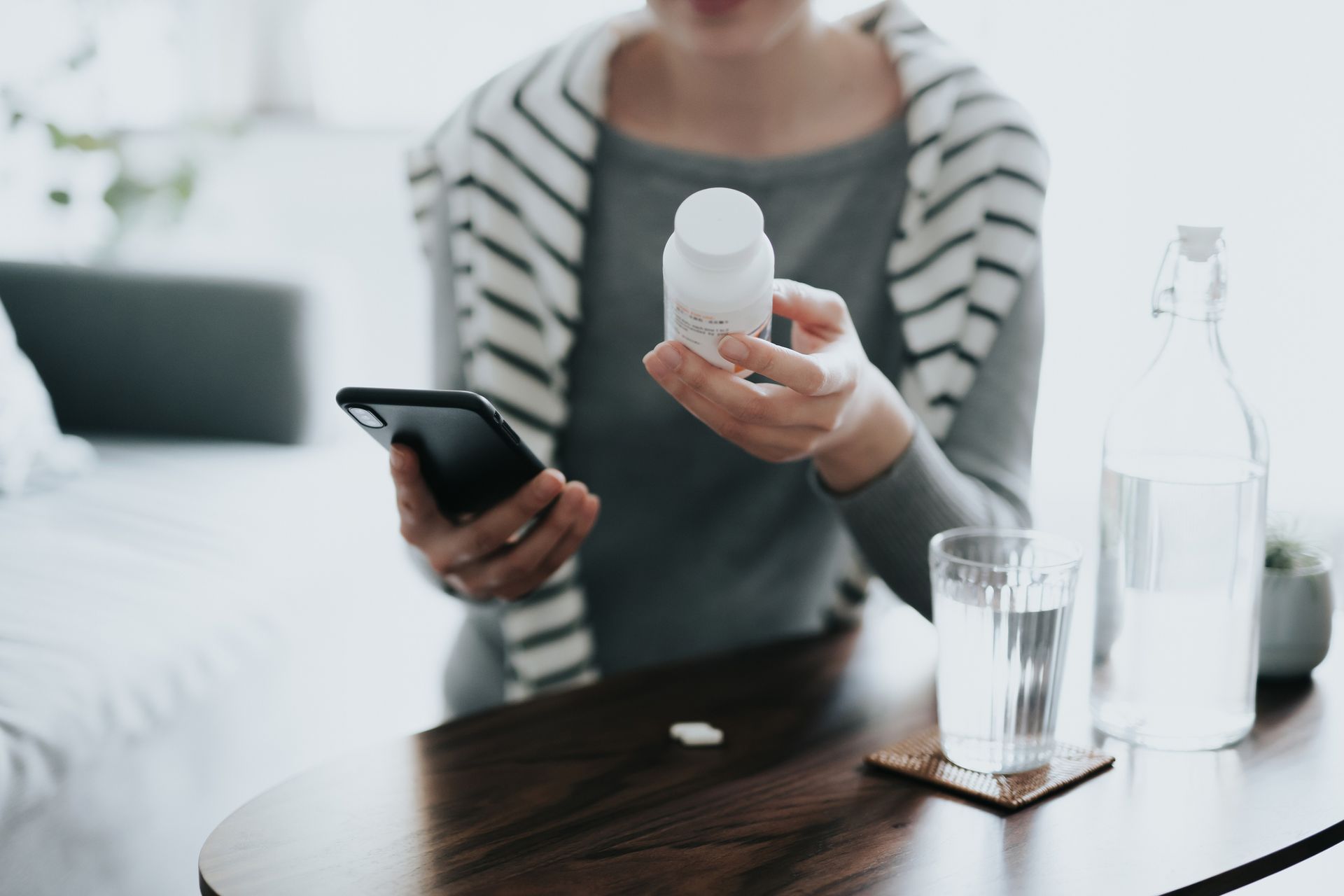 A woman is sitting at a table holding a bottle of pills and a cell phone.