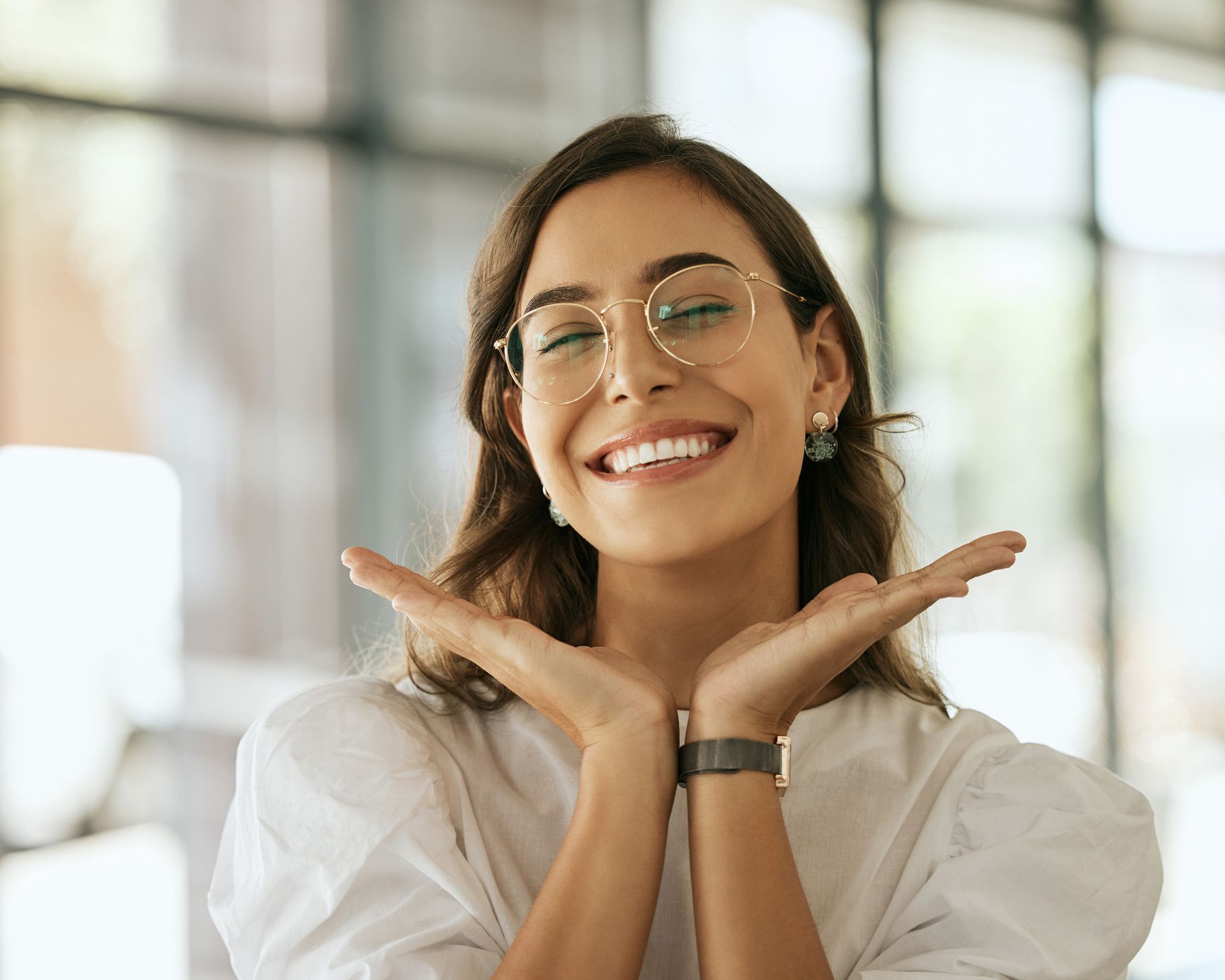 A woman wearing glasses is smiling with her hands on her face.