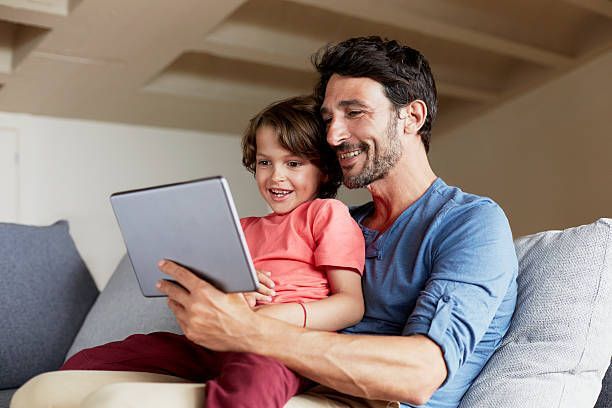 A man and a child are sitting on a couch looking at a tablet.