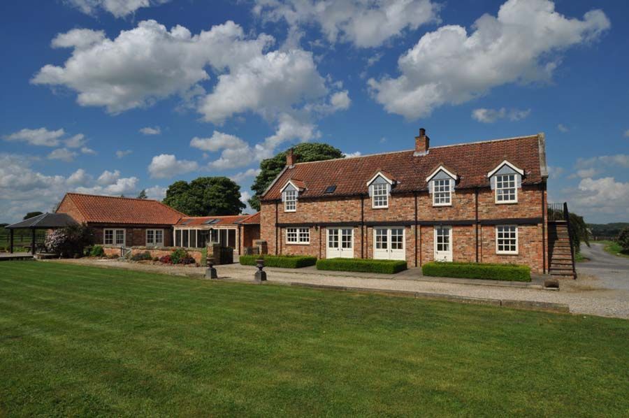 Brick country house with dormers and outbuilding on a green lawn, under a blue sky with fluffy clouds.