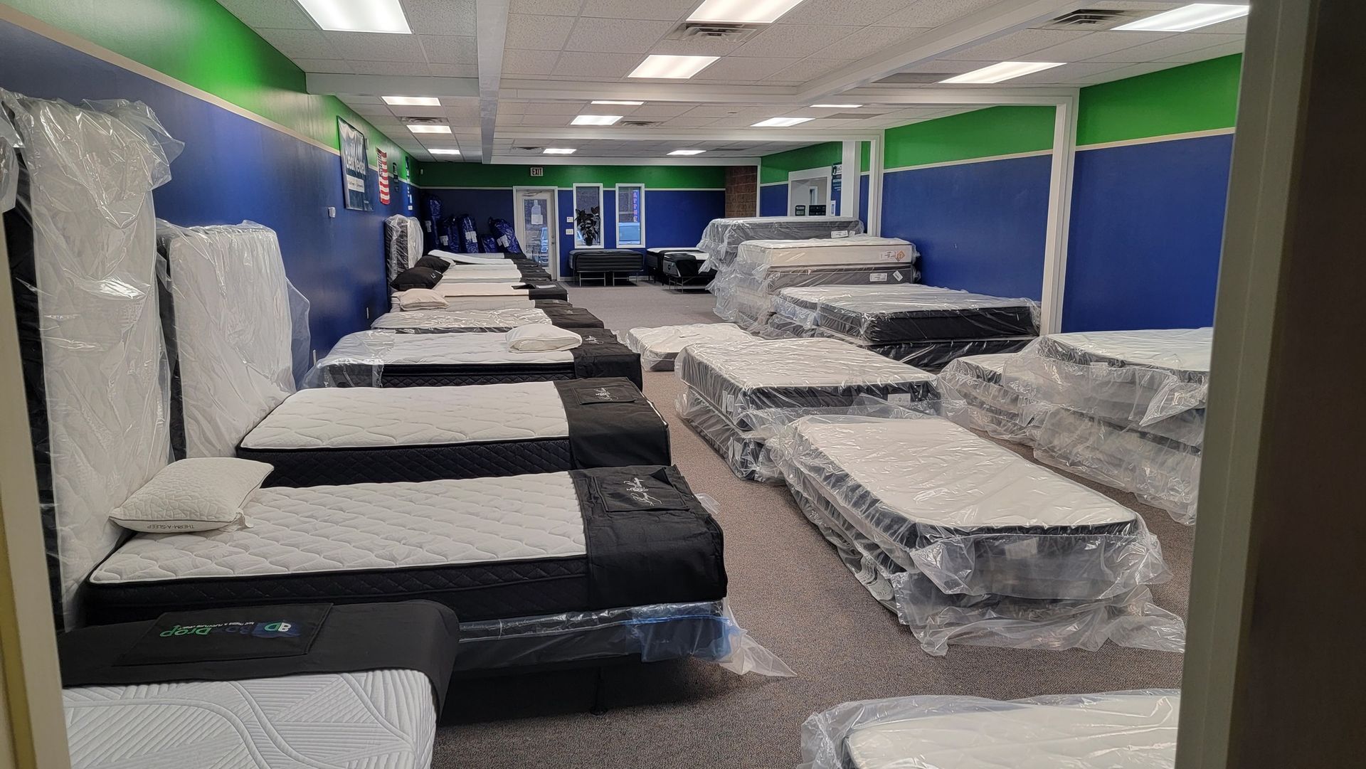 A brightly lit mattress store with numerous mattresses on display, some wrapped in plastic. Walls are blue and green.