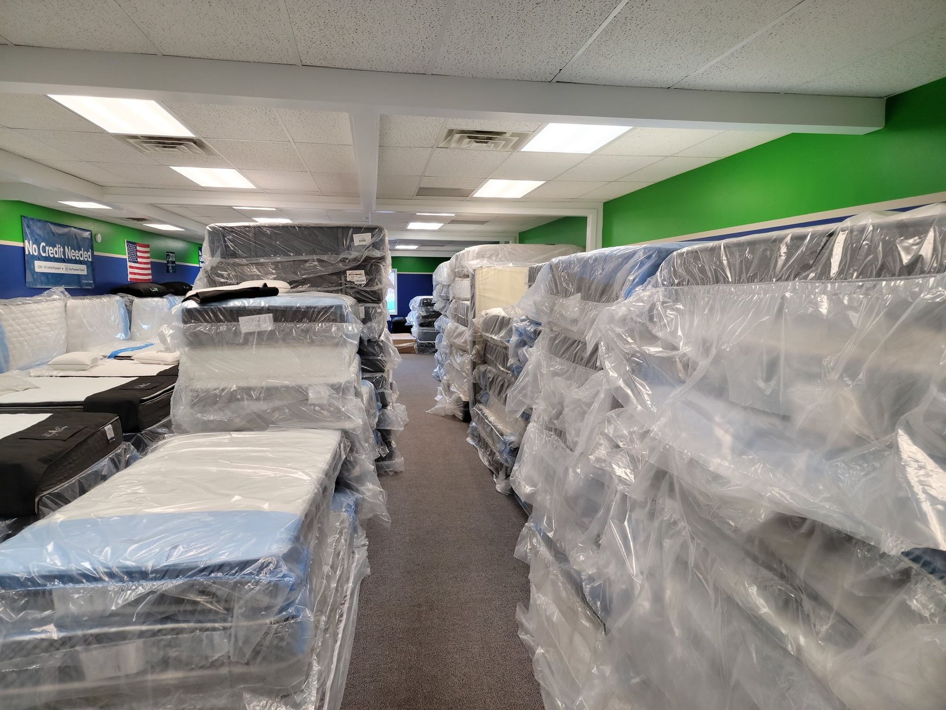Man with curly hair and painted nails reclines on a mattress in front of a brick wall, wearing a jacket, necklace, and black boots.