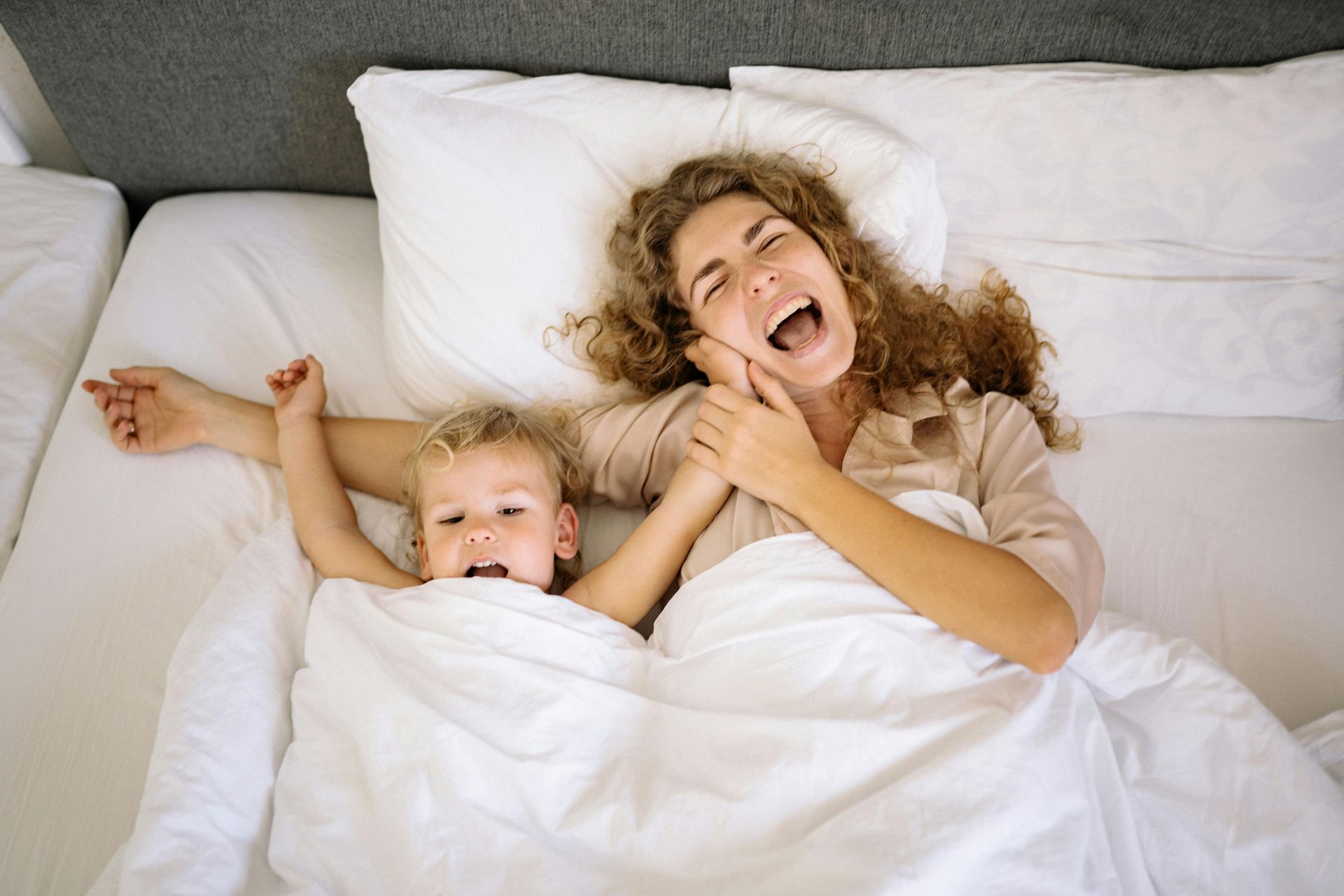 Woman and child laughing, lying in bed together under white covers.