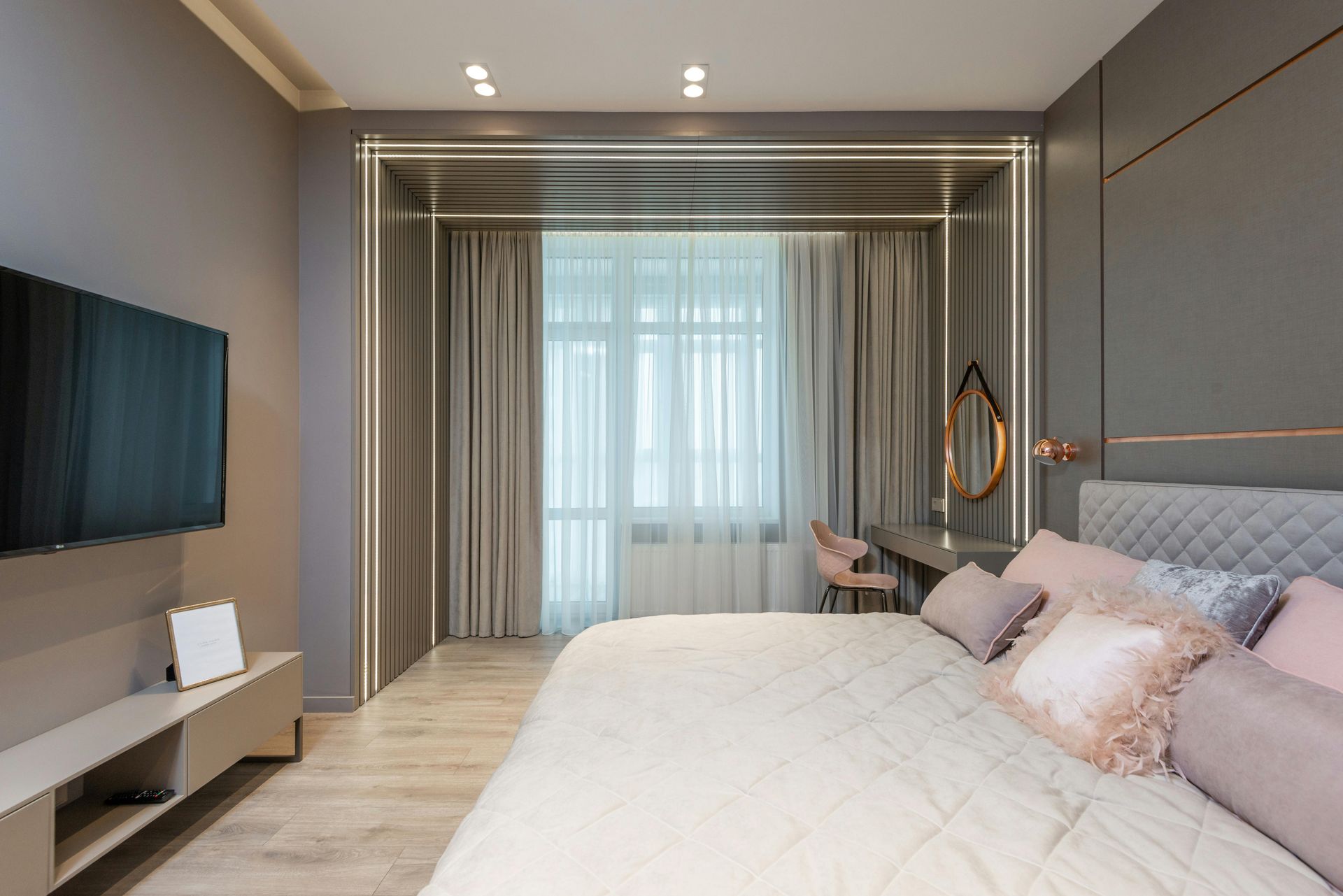 A modern bedroom with a bed, desk, TV, and window. Gray walls and bedding with rose-colored pillows and accents.