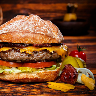 Ein Cheeseburger mit Speck, Tomaten, Salat und Essiggurken auf einem Holztisch, dazu eine Kirschtomate und Zwiebeln als Beilage.