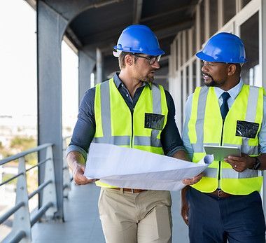 Two construction workers wearing hard hats and safety vests are looking at a blueprint.