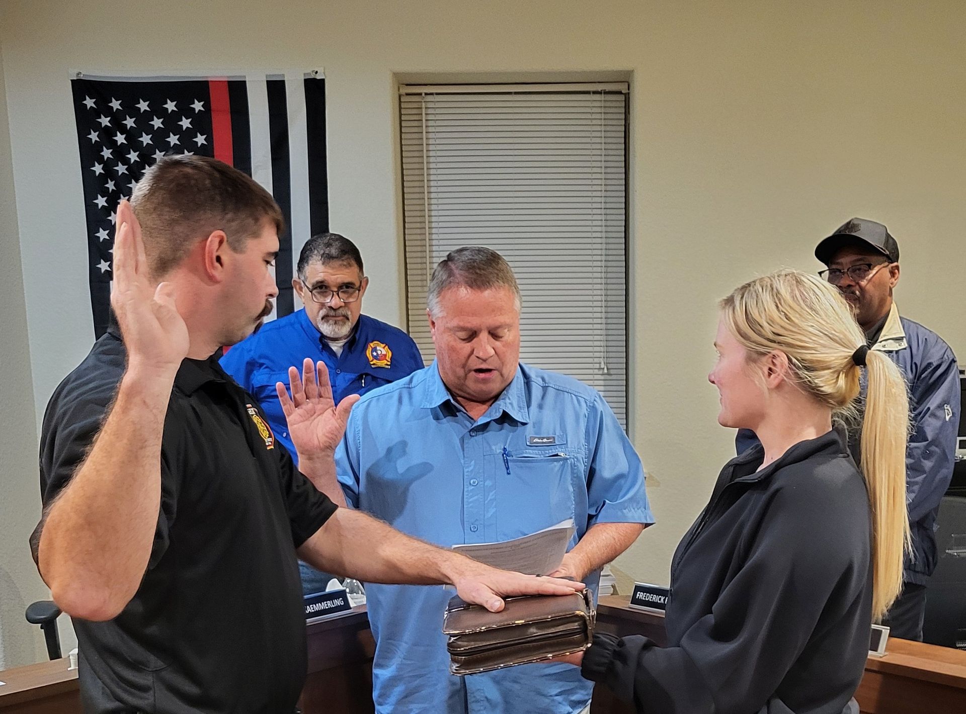Man being sworn in, raising hand, with others observing. Ceremony in a room with a flag.