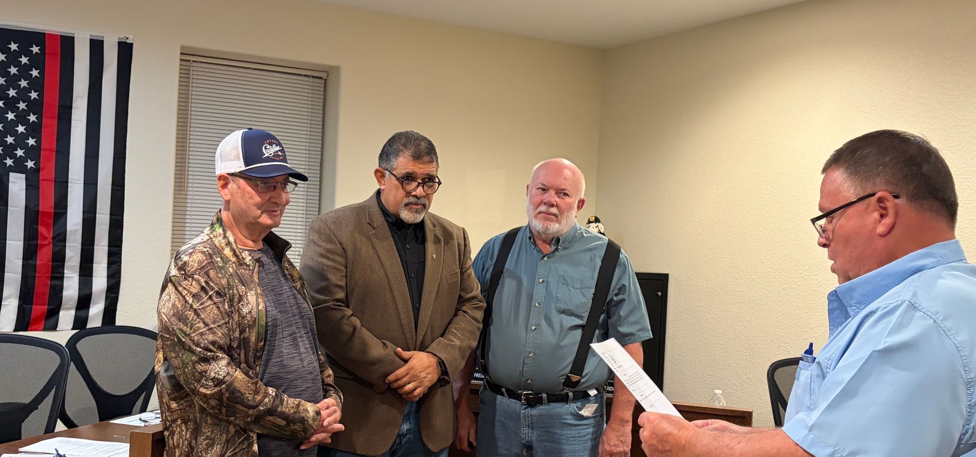 Four men in an office setting, one reading from a paper, three others watching. A thin red line flag hangs on the wall.