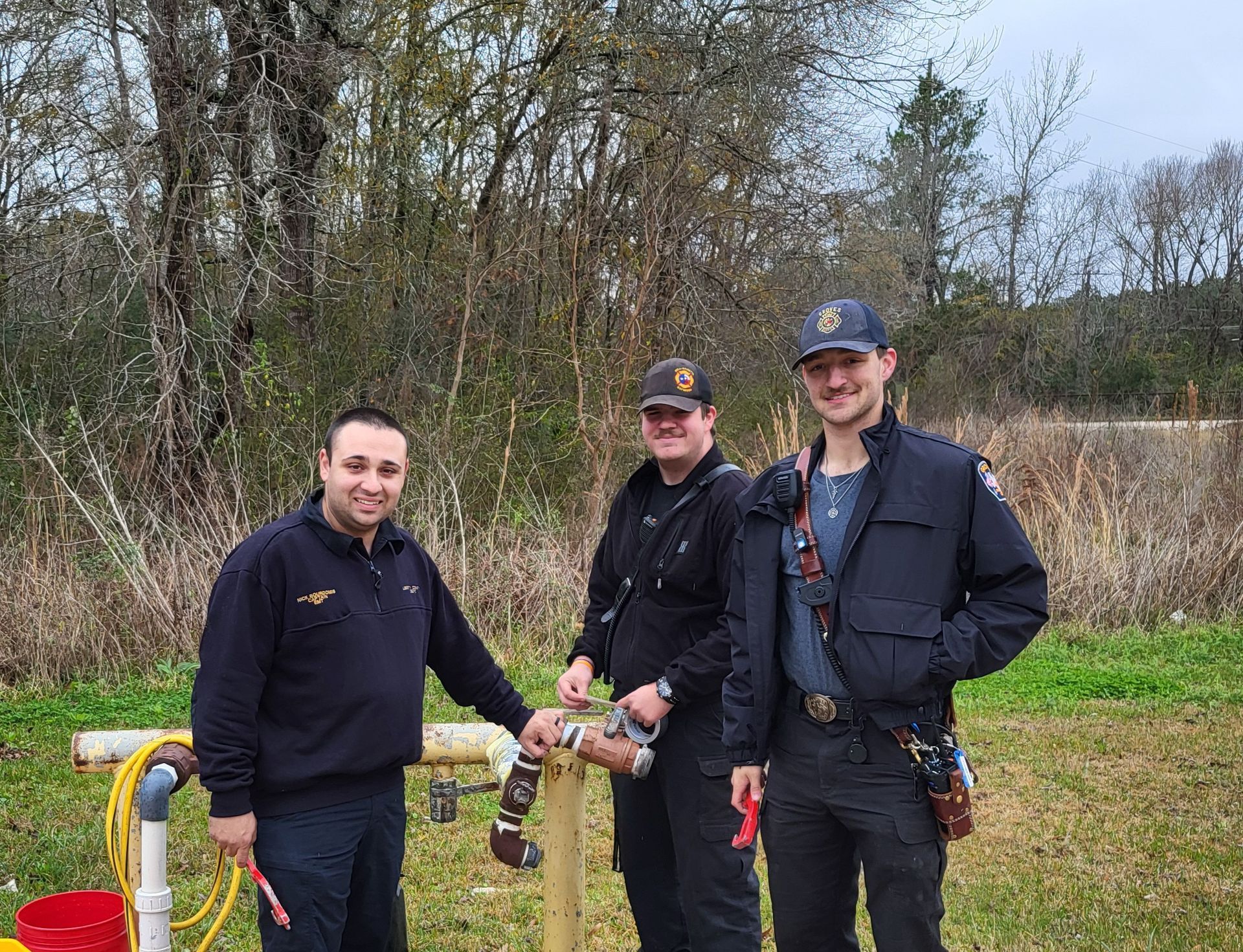 Three people in dark uniforms by a pipe structure, one holding a wrench, outdoors in a field.