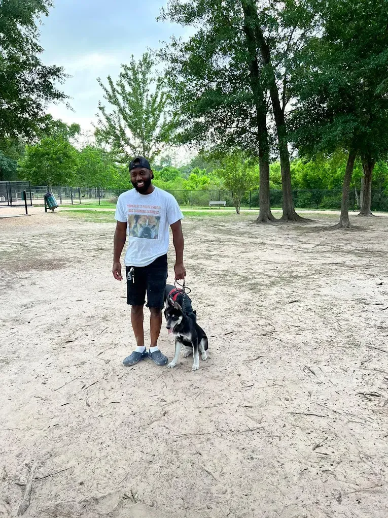 Man in shorts and t-shirt holds dog leash in a park with dog.