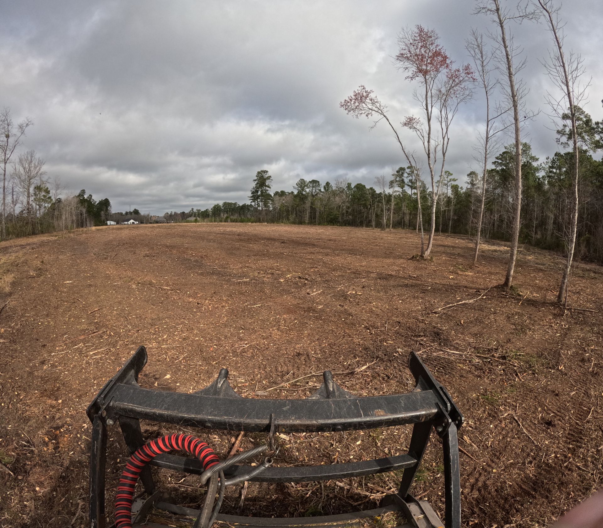 View from a skid-steer grapple attachment looking out over a cleared, woodchipped field under a cloudy sky.
