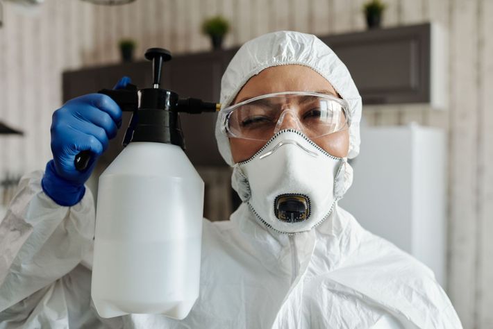 Une personne portant une combinaison de protection, un masque et des lunettes de protection, tient un flacon pulvérisateur à l'intérieur.