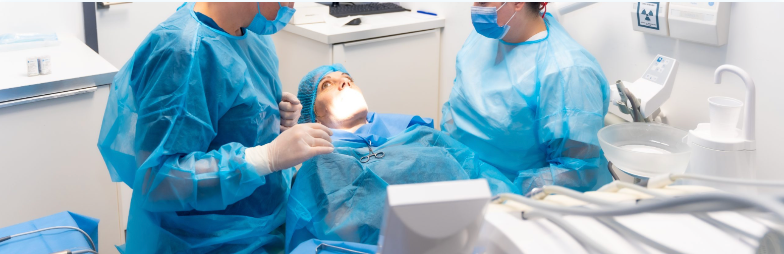 Two medical professionals in blue scrubs and masks attend to a patient in a dental chair.