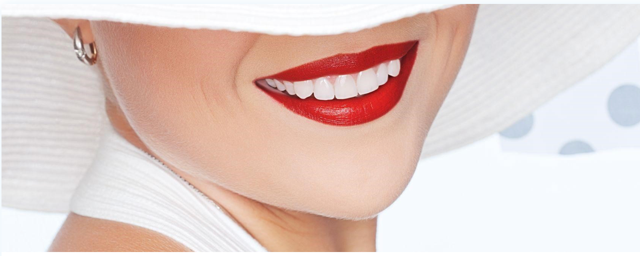 A person smiling widely, showing bright white teeth and red lipstick. Wearing a white hat and earring.