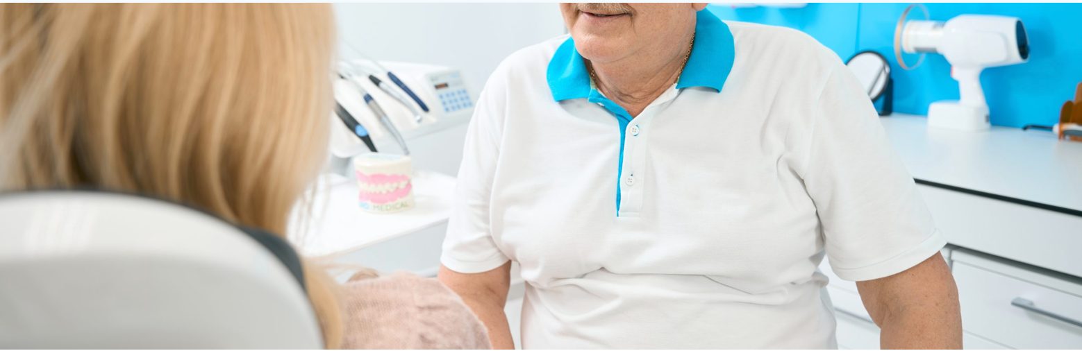 Person in a dentist's office wearing a white shirt with a teal collar; another person with blonde hair is sitting in the dentist's chair.