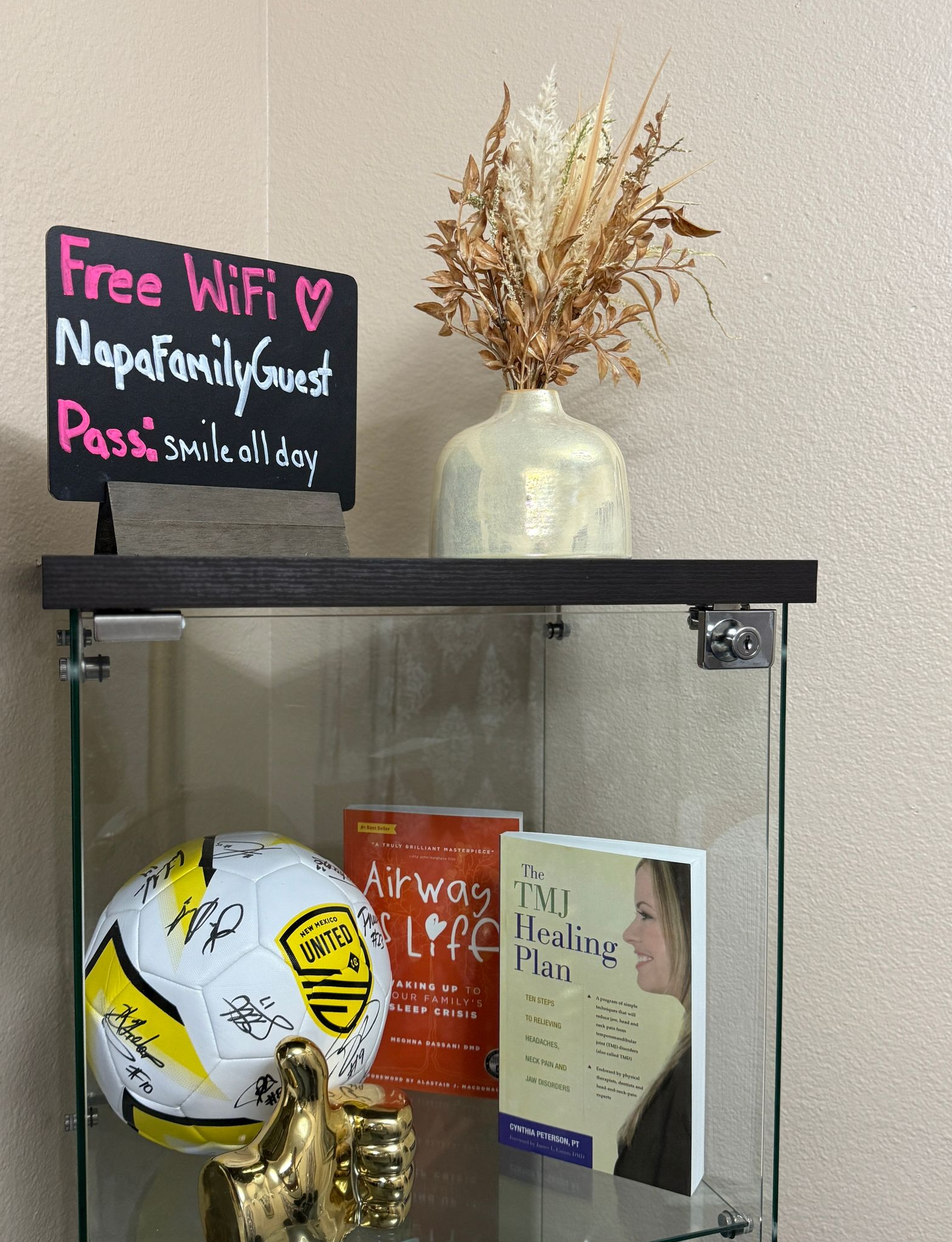 Glass display cabinet with books, soccer ball, and vase with dried flowers, topped by a chalkboard with WiFi information.