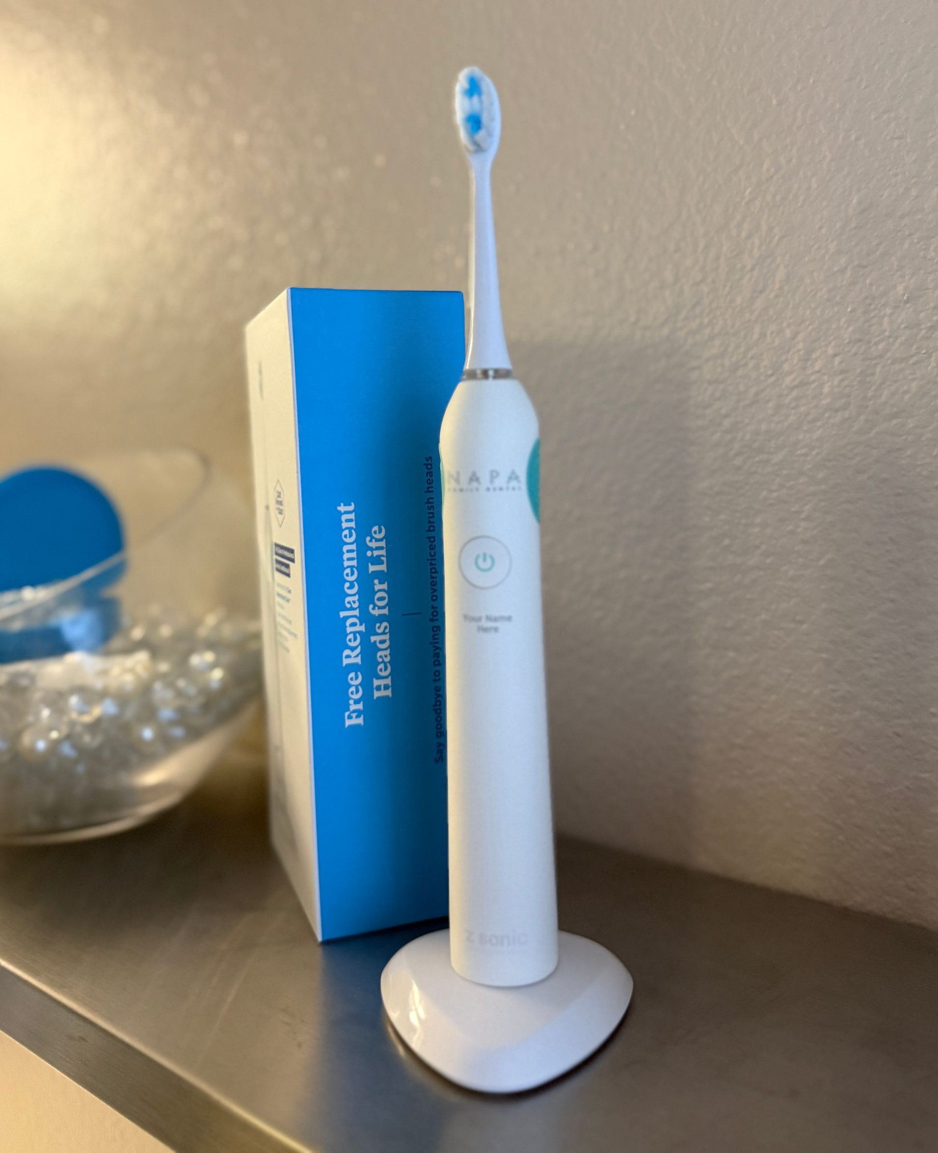 White electric toothbrush in a stand, next to its box, on a metal surface.