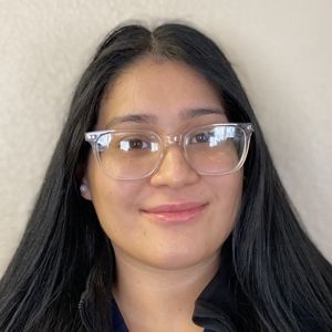 Person with long black hair, wearing clear glasses, smiling slightly, against a light wall.