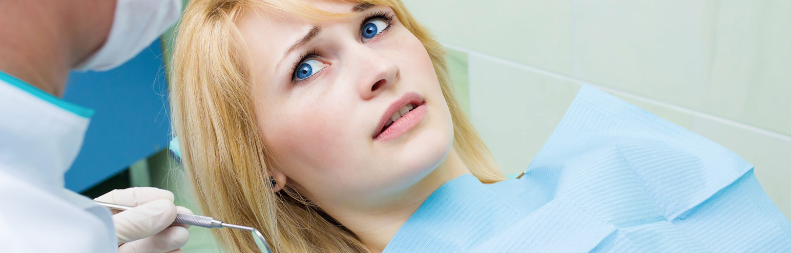 A woman with blonde hair is at the dentist, looking scared with wide eyes.