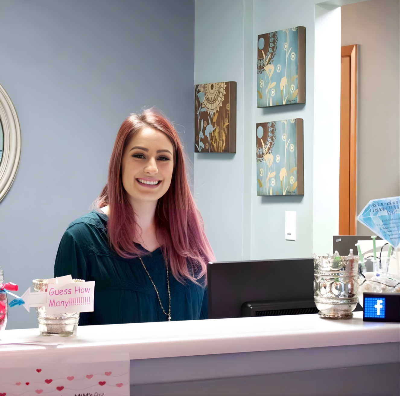 Woman with pink hair smiles at a reception desk. Artwork hangs on a light blue wall.