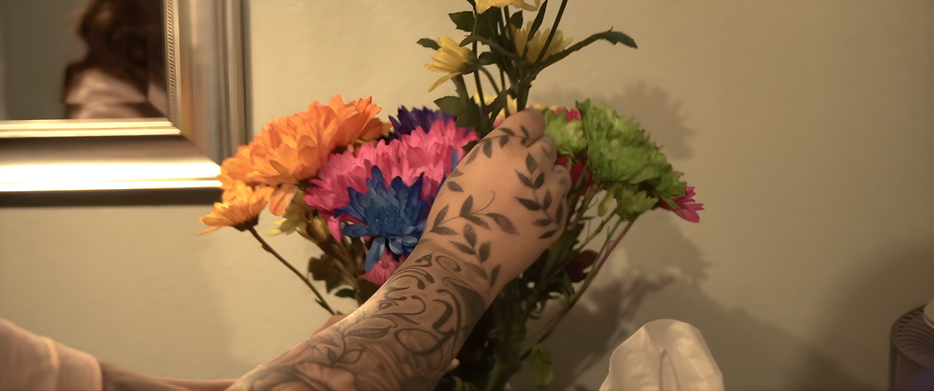 A person with arm tattoos arranging colorful flowers in a vase near a mirror.