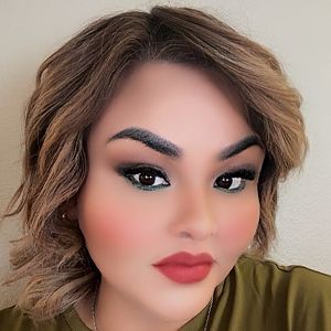 Woman with brown hair and full makeup, wearing a green top, looking at the camera.