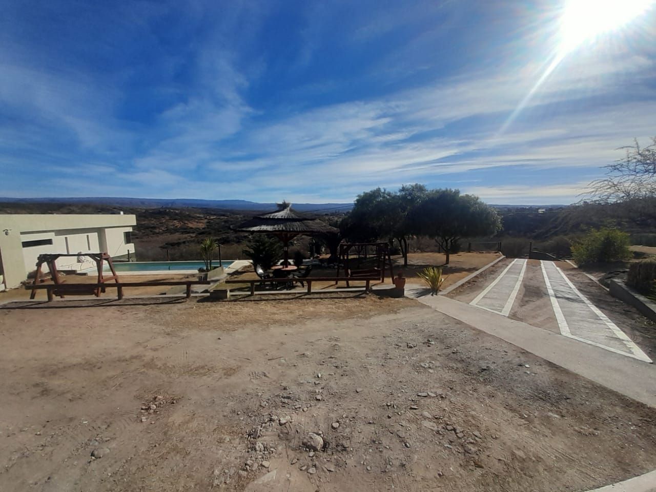 Una escena soleada al aire libre con un edificio, piscina, árboles y colinas distantes bajo un cielo azul.