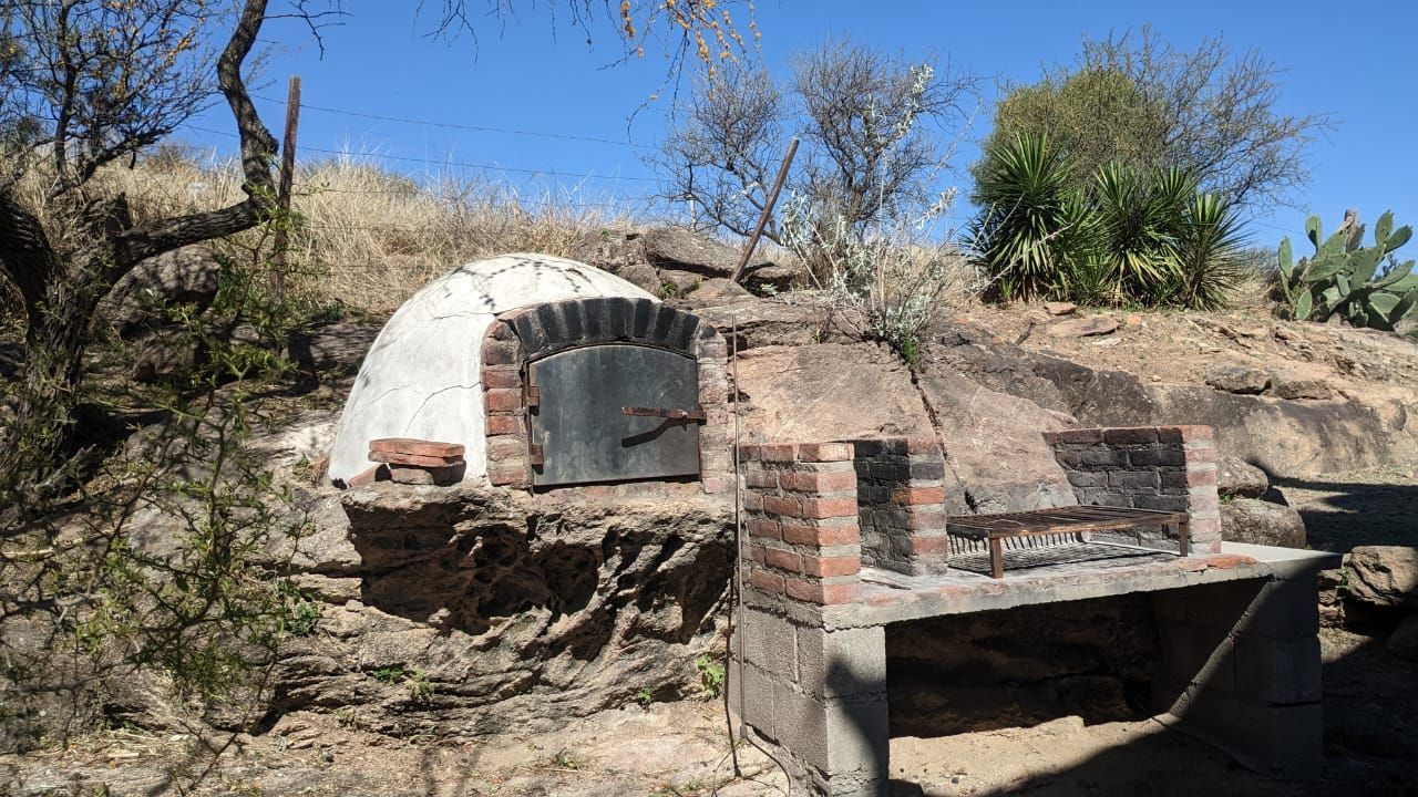 Horno de ladrillo y parrilla construidos en una ladera rocosa, rodeados de maleza seca y cactus bajo un cielo azul.