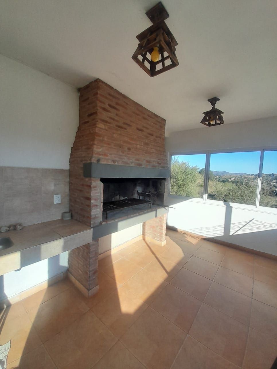 Parrilla de ladrillo en una habitación iluminada por el sol con vista al exterior, dos artefactos de iluminación de madera y pisos de baldosas.