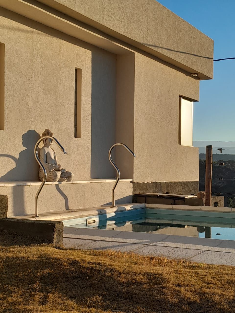 Una persona sentada en una silla junto a una piscina al lado de un edificio, bañada por la luz del sol.