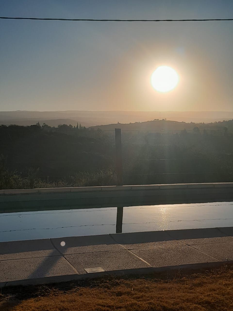 Puesta de sol sobre una línea de árboles distante reflejada en el agua, con una valla en primer plano.