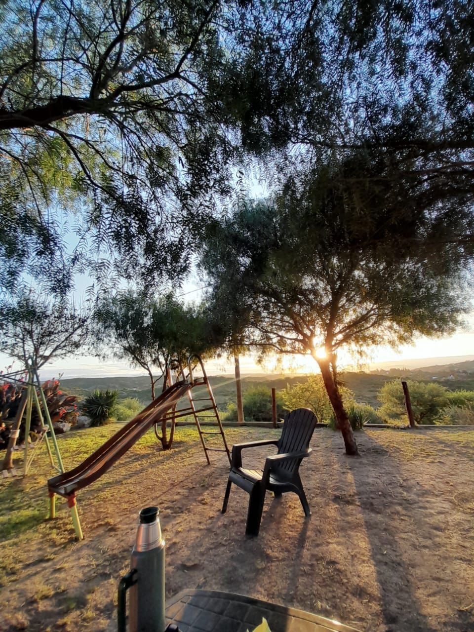 Parque infantil con tobogán, columpios y sillas bajo los árboles al atardecer.