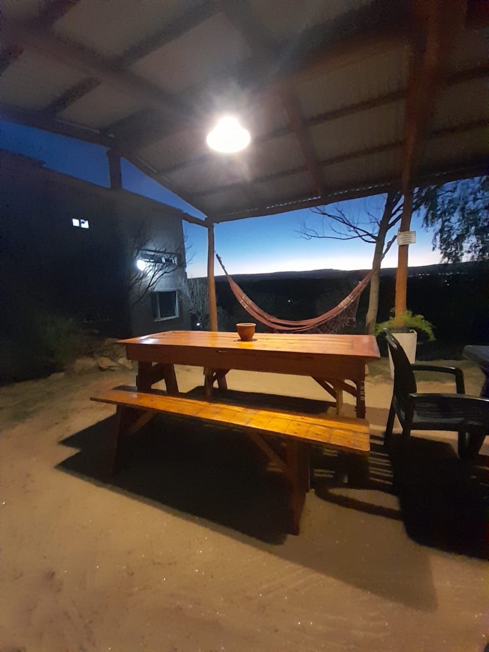 Patio exterior cubierto al anochecer con mesa de madera, banco, hamaca y montañas a lo lejos.