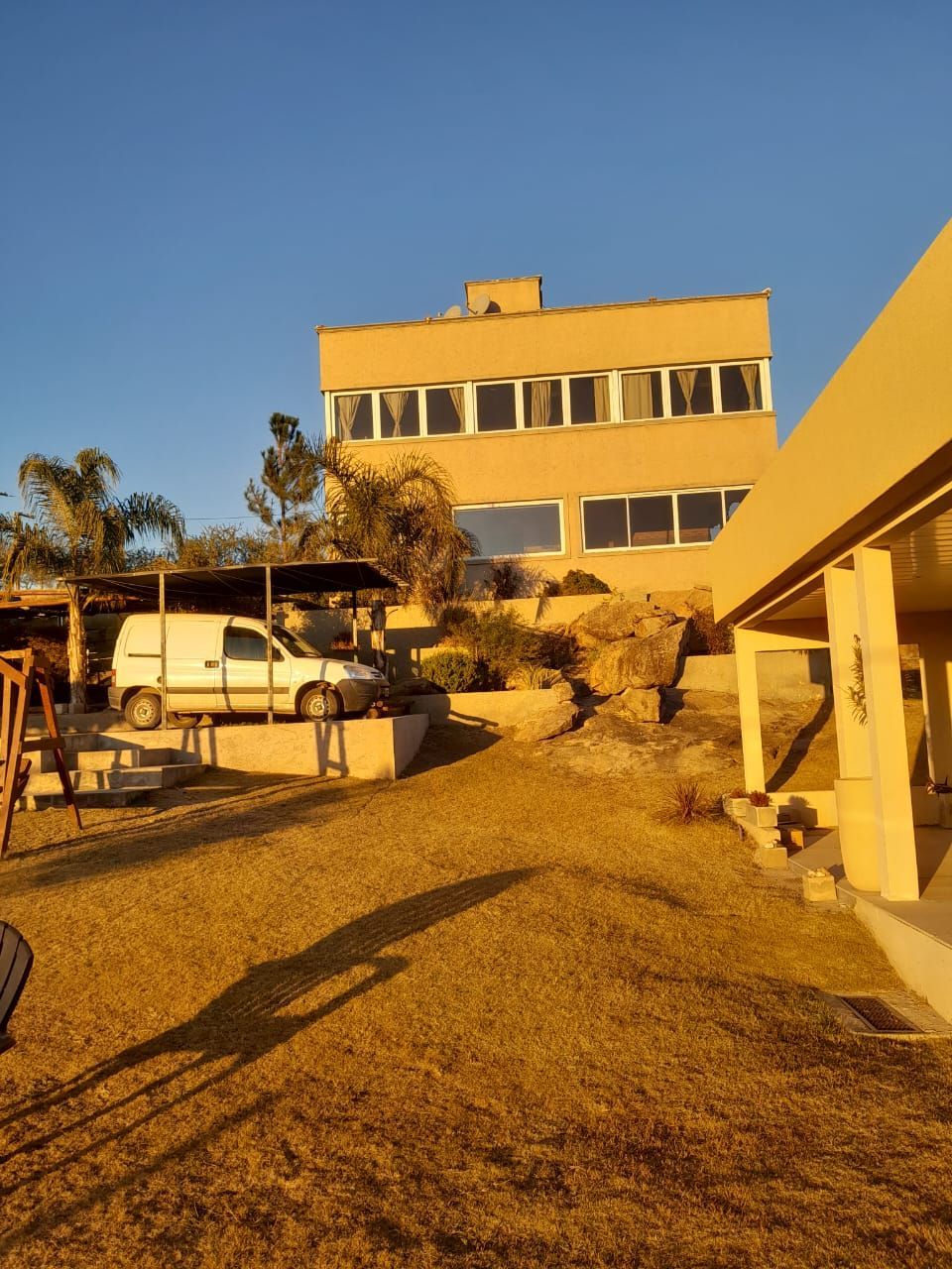 Un edificio de tres pisos de color canela con una camioneta estacionada afuera; patio con césped; cielo brillante.