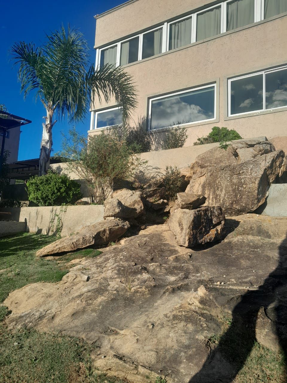 Casa color canela con grandes ventanales, paisaje rocoso y palmera contra un cielo azul.