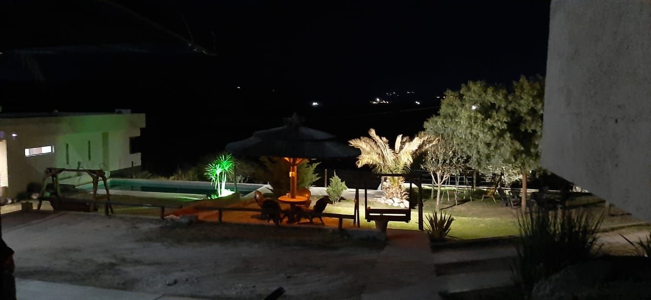 Escena nocturna al aire libre con cenador iluminado, asientos y árboles. Vista de una casa y piscina al fondo.