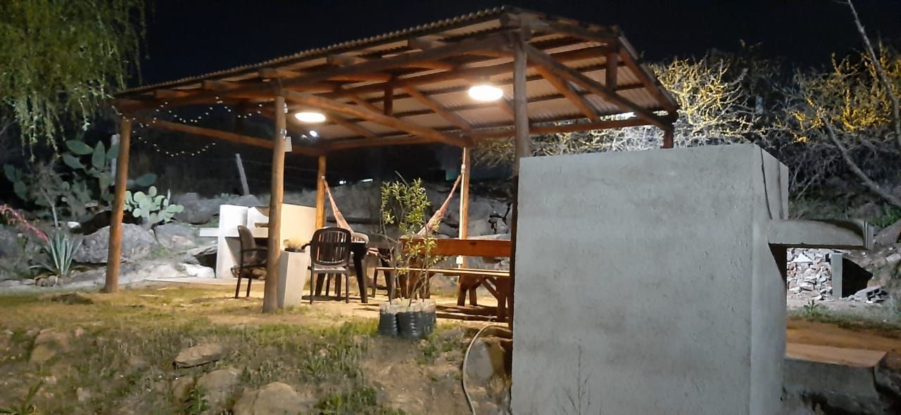 Zona de estar al aire libre por la noche bajo un dosel de madera con mesas y sillas iluminadas por luces de techo.