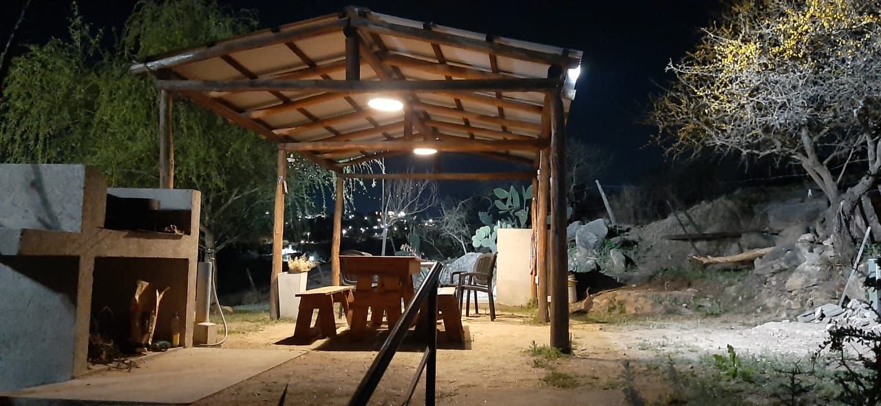 Un comedor exterior cubierto e iluminado por la noche, con parrilla, mesa, sillas y árboles circundantes.