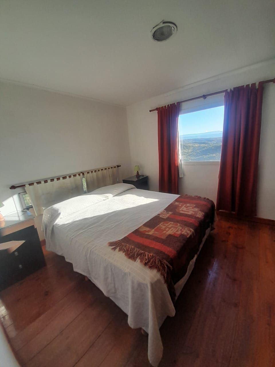 Dormitorio con cama, cortinas rojas, suelo de madera y ventana con vistas a un paisaje.