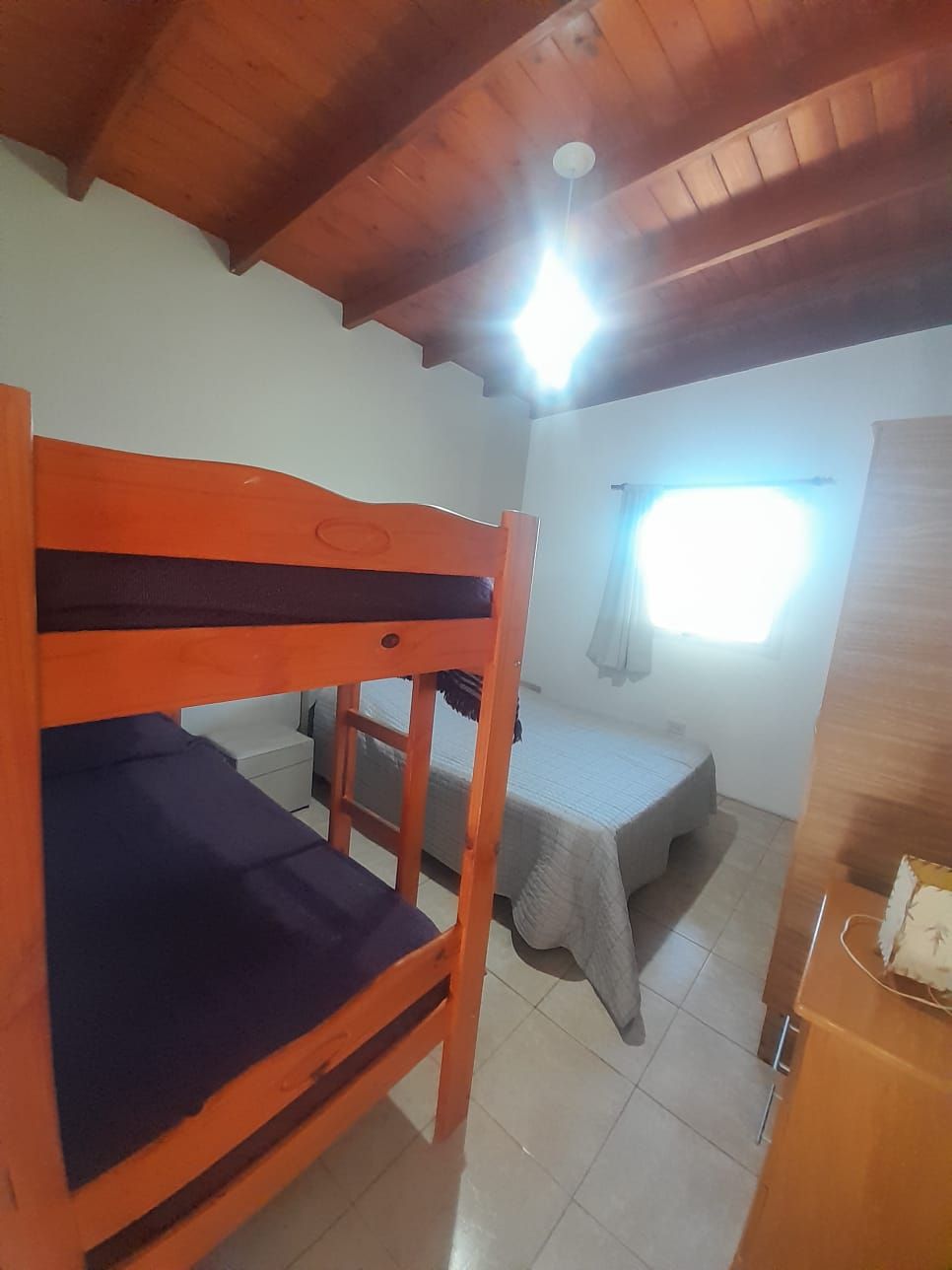 Litera en una habitación con cama, ventana y techo de madera. Luz brillante.