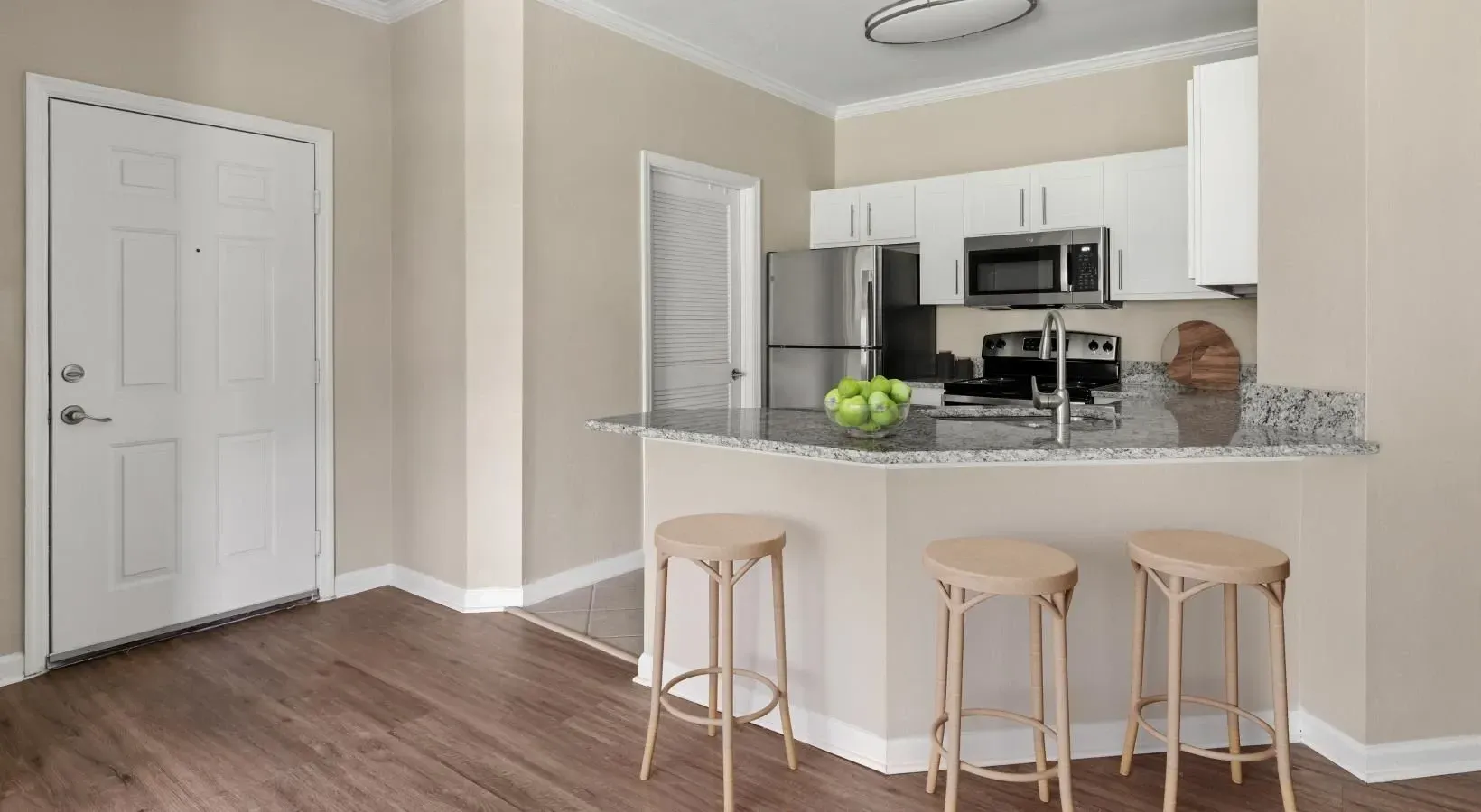 An open-concept apartment interior with wood flooring, a white front door, and a kitchen featuring white cabinets.