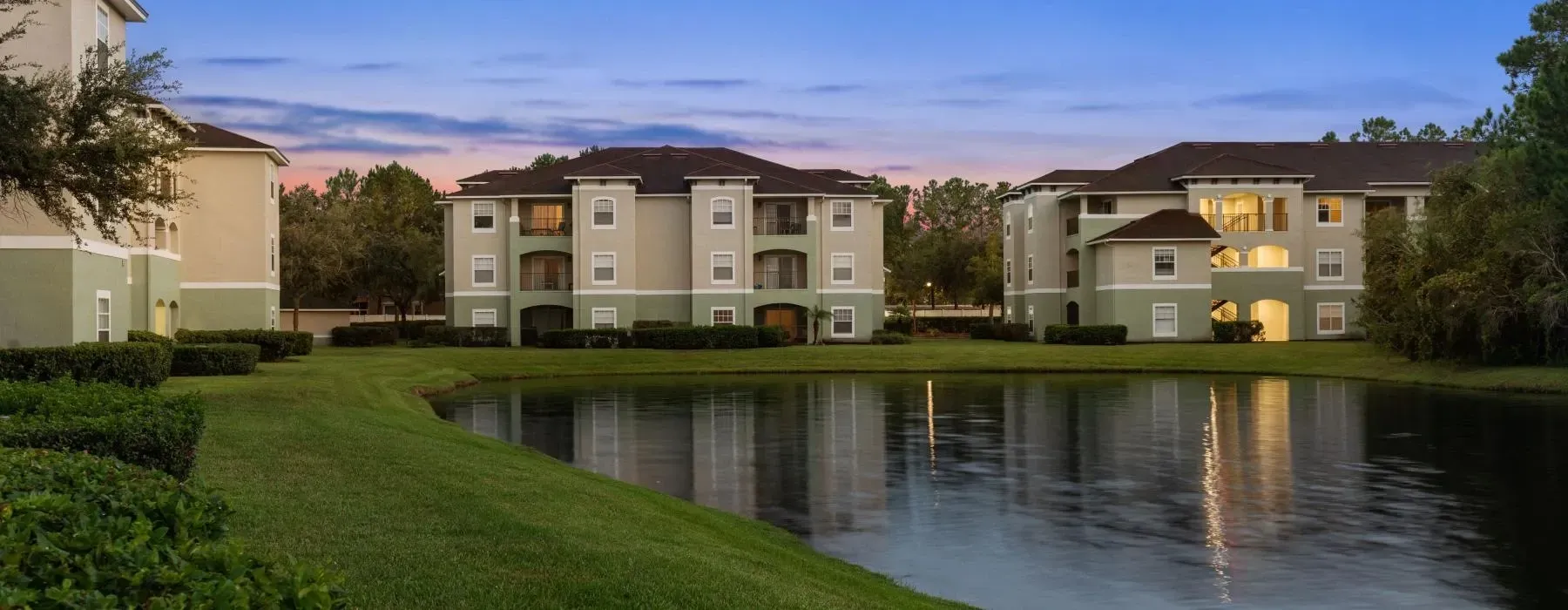 Three multi-story apartment buildings with tan and green siding sit behind a pond at sunset at The Vale at Baymeadows.
