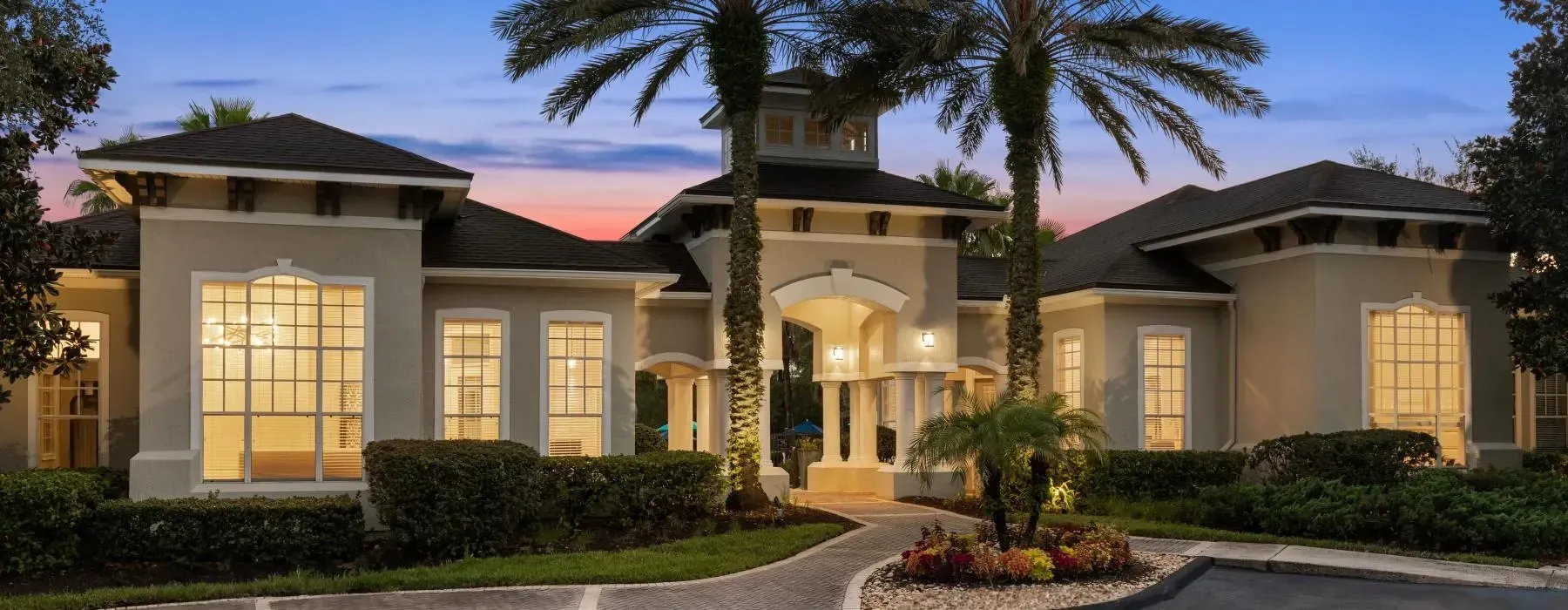 A tan, luxury residential building with a central cupola, lit windows, and palm trees at twilight.